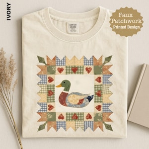 May include: An ivory t-shirt featuring a central faux patchwork printed design of a mallard duck surrounded by a border of small red hearts and various patterned fabric squares in shades of blue, green, tan, and cream with a crew neckline.