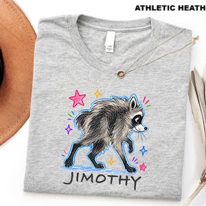 May include: An athletic heather gray t-shirt featuring a graphic of a furry raccoon with black and white markings. The raccoon is surrounded by colorful hand-drawn style stars and sparkles. The name JIMOTHY is printed in black below the raccoon.