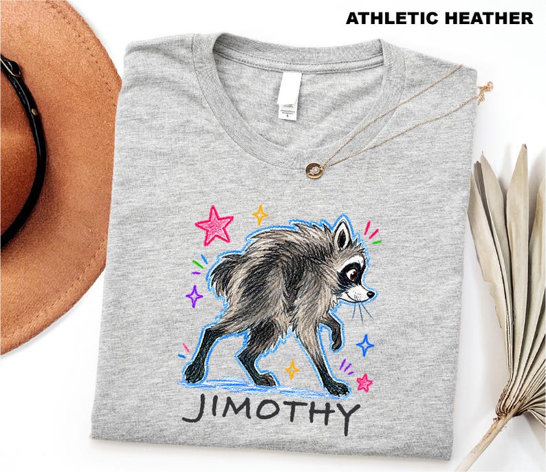 May include: An athletic heather gray t-shirt featuring a graphic of a furry raccoon with black and white markings. The raccoon is surrounded by colorful hand-drawn style stars and sparkles. The name JIMOTHY is printed in black below the raccoon.