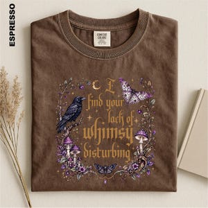 Puede incluir: Una camiseta de cuello redondo color marr&oacute;n espresso con un estampado gr&aacute;fico de un cuervo negro, setas moradas y mariposas. El texto dice I find your lack of whimsy disturbing, rodeado de elementos florales, estrellas y una luna creciente.