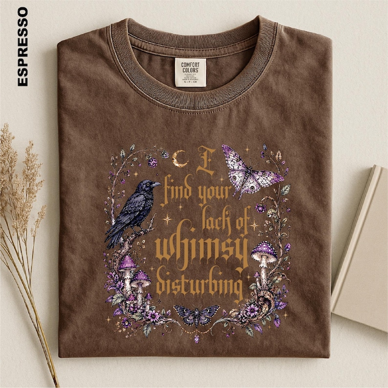 Puede incluir: Una camiseta de cuello redondo color marr&oacute;n espresso con un estampado gr&aacute;fico de un cuervo negro, setas moradas y mariposas. El texto dice I find your lack of whimsy disturbing, rodeado de elementos florales, estrellas y una luna creciente.