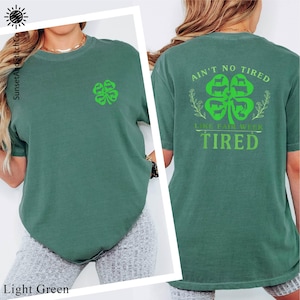 May include: Light green t-shirt with a small green shamrock on the front and a larger shamrock design on the back. The back design includes the text "Ain't No Tired Like Fair Week Tired".