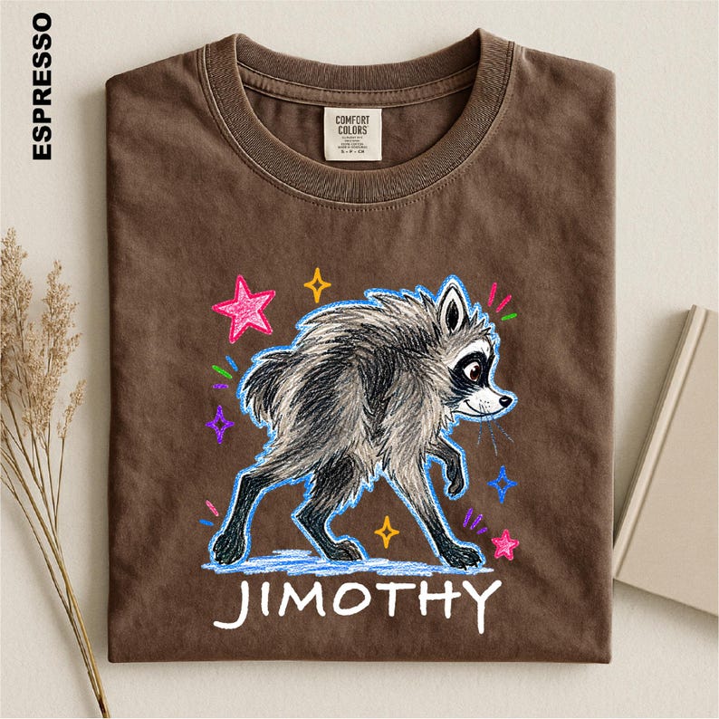 May include: An espresso brown t-shirt featuring a graphic of a grey raccoon with a sketch-like texture. The raccoon is surrounded by colorful hand-drawn style stars in pink, yellow, and blue. The name JIMOTHY is printed in white below the raccoon.