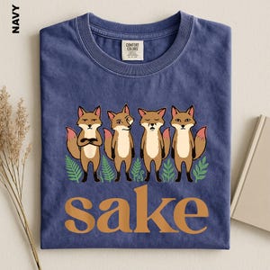 May include: A navy blue crew neck t-shirt featuring a graphic of four cartoon foxes standing above the word "sake" in large tan letters. The foxes are depicted with various expressions, surrounded by small green fern illustrations.