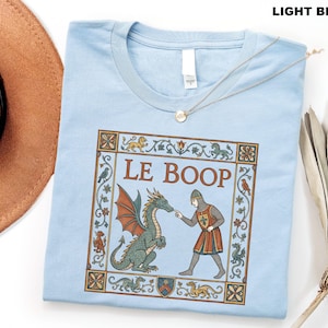 May include: A light blue t-shirt featuring a medieval-style graphic print of a knight touching a dragon's nose. The text LE BOOP is centered above the figures, surrounded by a decorative border with small dragons, birds, and floral motifs.