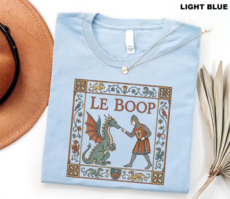 May include: A light blue t-shirt featuring a medieval-style graphic print of a knight touching a dragon's nose. The text LE BOOP is centered above the figures, surrounded by a decorative border with small dragons, birds, and floral motifs.