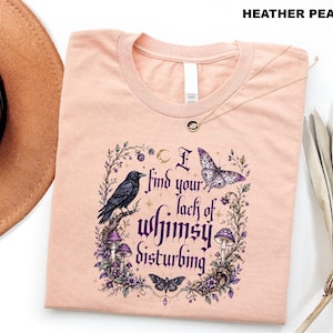 Puede incluir: Una camiseta color melocot&oacute;n jaspeado con un estampado gr&aacute;fico que incluye el texto "I find your lack of whimsy disturbing". El dise&ntilde;o muestra un cuervo negro, una mariposa morada, una polilla y setas rodeadas de enredaderas y una luna.