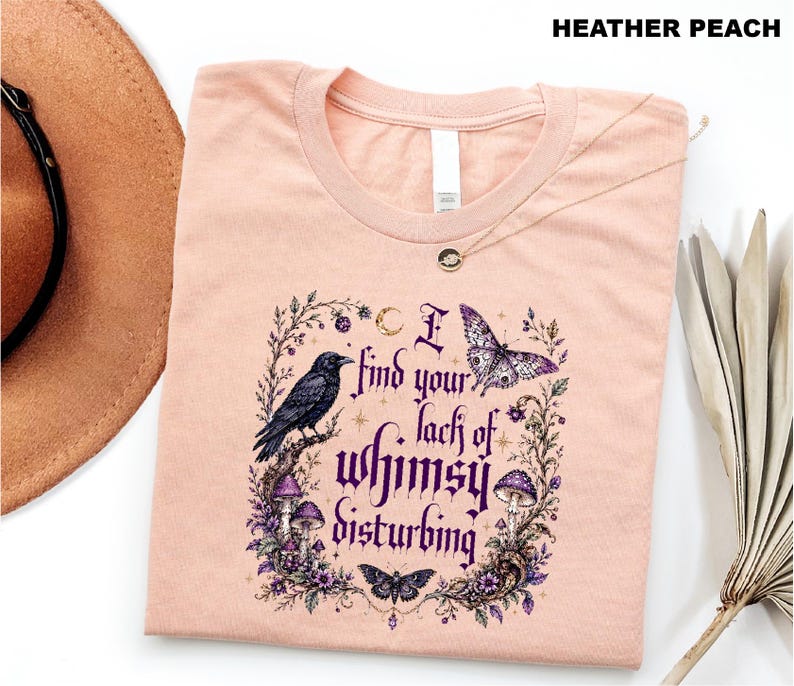 Puede incluir: Una camiseta color melocot&oacute;n jaspeado con un estampado gr&aacute;fico que incluye el texto "I find your lack of whimsy disturbing". El dise&ntilde;o muestra un cuervo negro, una mariposa morada, una polilla y setas rodeadas de enredaderas y una luna.