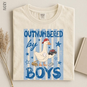 A t-shirt with a graphic design featuring a cartoon goose holding a baseball and a toy truck, along with the text "OUTNUMBERED BY BOYS".