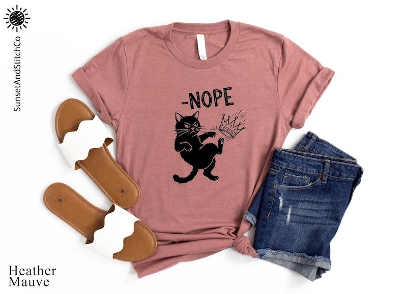 May include: Heather mauve t-shirt with a black graphic of a cat kicking a crown with the word '-NOPE' above it. The shirt is paired with denim shorts and white sandals with a wavy strap design. The SunsetAndStitchCo logo is in the top left corner.