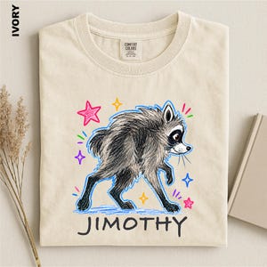 May include: An ivory-colored t-shirt featuring a graphic of a gray raccoon drawn in a crayon style. The raccoon is surrounded by colorful hand-drawn stars and small marks. Below the raccoon, the name JIMOTHY is printed in black capital letters.