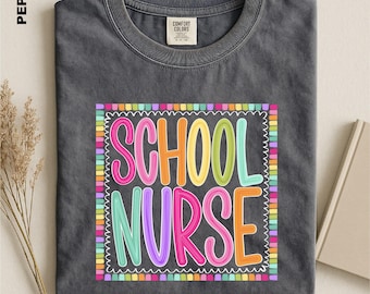 Pode incluir: Uma t-shirt de gola redonda cinzento-carvão com uma impressão gráfica colorida com o texto SCHOOL NURSE numa fonte lúdica e multicolorida. O design está inserido numa borda quadrada feita de pequenos blocos retangulares vibrantes.