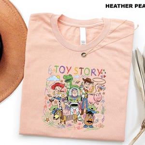 A pink t-shirt with a colorful graphic of characters from the Toy Story movie, including Woody, Buzz Lightyear, and other beloved characters, along with various other elements.