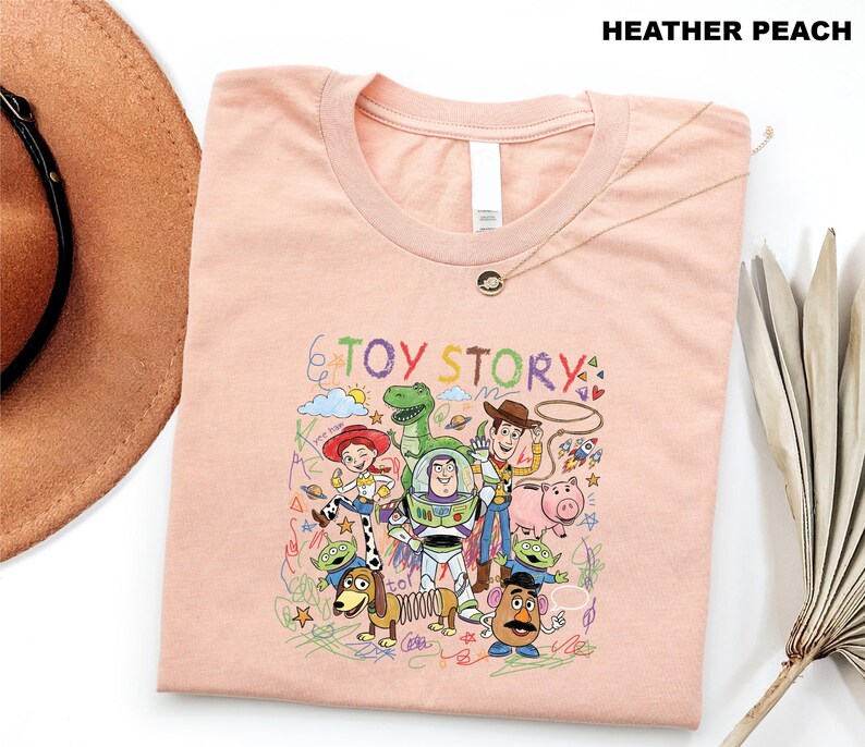 A pink t-shirt with a colorful graphic of characters from the Toy Story movie, including Woody, Buzz Lightyear, and other beloved characters, along with various other elements.
