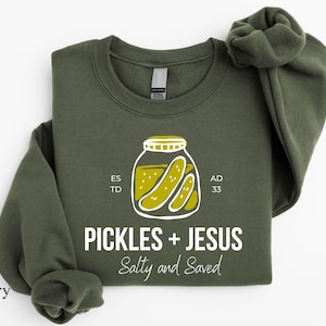 A military green sweatshirt with a graphic design featuring a jar of pickles and the text "Pickles + Jesus Salty and Saved" printed on it.