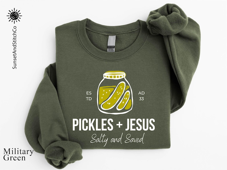 A military green sweatshirt with a graphic design featuring a jar of pickles and the text "Pickles + Jesus Salty and Saved" printed on it.