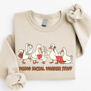 May include: Sand-colored sweatshirt featuring a cartoon illustration of five geese. The geese are depicted in various social worker roles, carrying a briefcase, holding a phone, and using a laptop. The text "DOING SOCIAL WORKER STUFF" is printed below the geese.