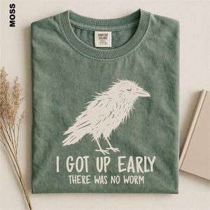 May include: A moss green crew neck t-shirt featuring a white graphic illustration of a bird. The text "I GOT UP EARLY THERE WAS NO WORM" is printed in white below the bird. The shirt has a Comfort Colors brand label inside the collar.