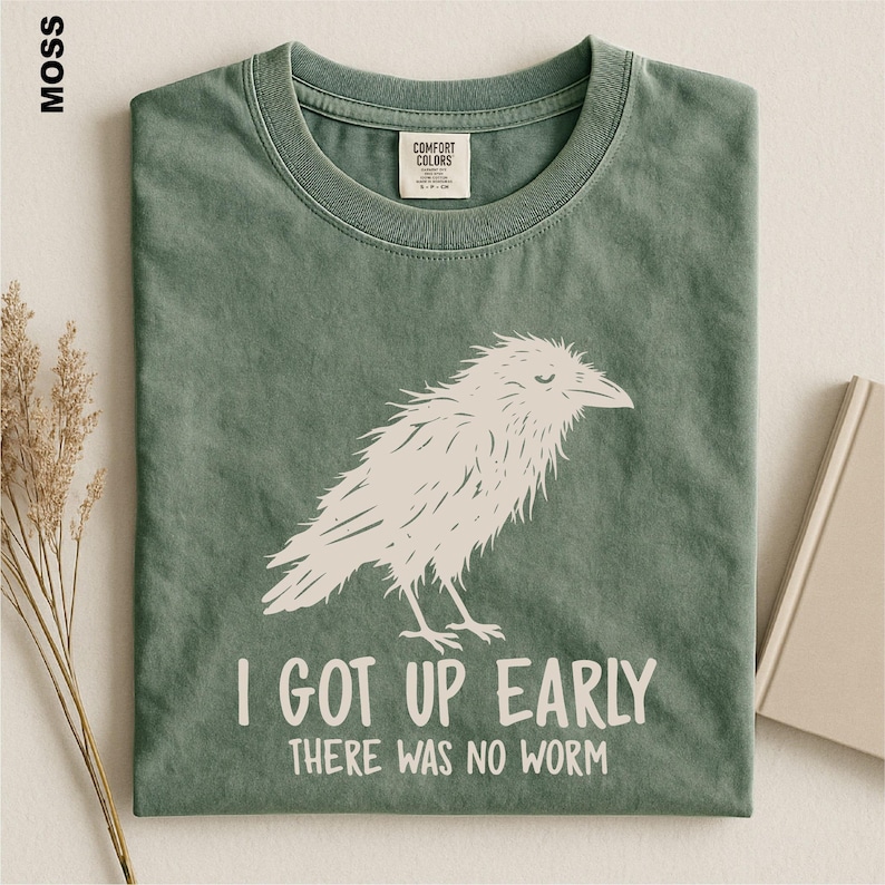 May include: A moss green crew neck t-shirt featuring a white graphic illustration of a bird. The text "I GOT UP EARLY THERE WAS NO WORM" is printed in white below the bird. The shirt has a Comfort Colors brand label inside the collar.