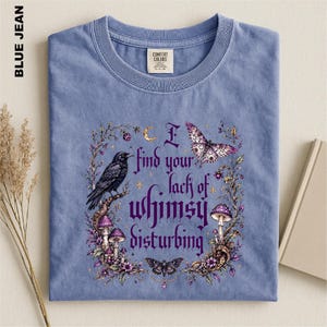Puede incluir: Una camiseta azul de cuello redondo con un estampado gr&aacute;fico que incluye el texto I find your lack of whimsy disturbing. El dise&ntilde;o presenta un cuervo negro, setas moradas, una polilla, una mariposa y elementos florales en un marco.