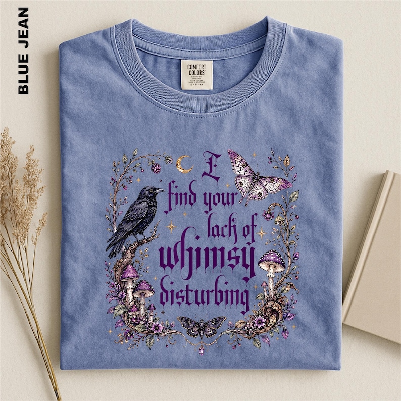 Puede incluir: Una camiseta azul de cuello redondo con un estampado gr&aacute;fico que incluye el texto I find your lack of whimsy disturbing. El dise&ntilde;o presenta un cuervo negro, setas moradas, una polilla, una mariposa y elementos florales en un marco.