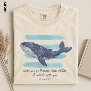 May include: An ivory t-shirt featuring a blue whale illustration centered over horizontal light blue brushstrokes. Below the whale, the text reads: when you go through deep waters, I will be with you Isaiah 43:2. The shirt has a crew neckline.