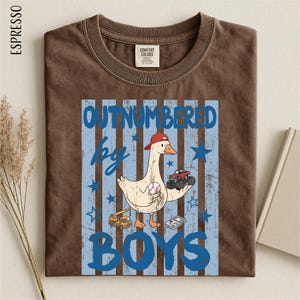 A brown t-shirt with a graphic design featuring a cartoon duck holding a baseball and a truck, along with the text "OUTNumbered by BOYS".