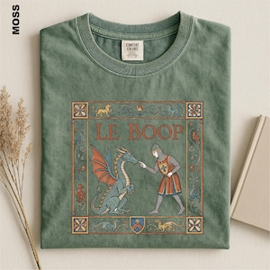 May include: A moss green crew neck t-shirt featuring a graphic print of a medieval-style knight touching the nose of a dragon. The text LE BOOP is printed above the figures, surrounded by a decorative border with floral and animal motifs.