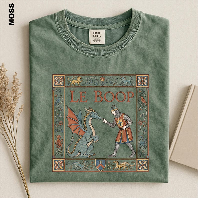 May include: A moss green crew neck t-shirt featuring a graphic print of a medieval-style knight touching the nose of a dragon. The text LE BOOP is printed above the figures, surrounded by a decorative border with floral and animal motifs.