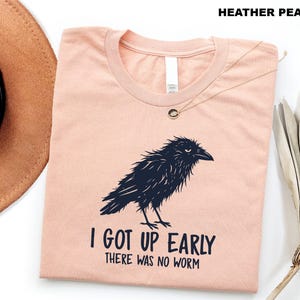 May include: A heather peach crew neck t-shirt featuring a dark navy blue graphic of a ruffled crow. Below the bird, the text reads I GOT UP EARLY THERE WAS NO WORM in a bold, dark navy blue font. The shirt is displayed flat with a round neckline.