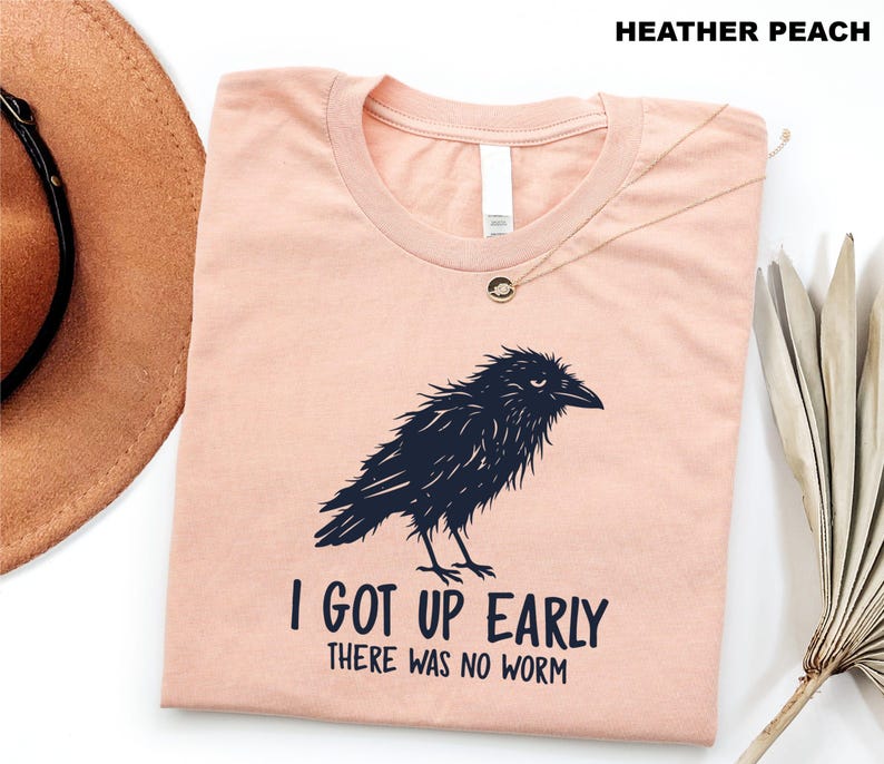 May include: A heather peach crew neck t-shirt featuring a dark navy blue graphic of a ruffled crow. Below the bird, the text reads I GOT UP EARLY THERE WAS NO WORM in a bold, dark navy blue font. The shirt is displayed flat with a round neckline.