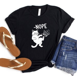 May include: Black t-shirt featuring a white cat graphic kicking a crown, with the word "-NOPE" above. The shirt is styled with denim shorts and brown sandals with gold straps. The brand name "SunsetAndStitchCo" is visible.