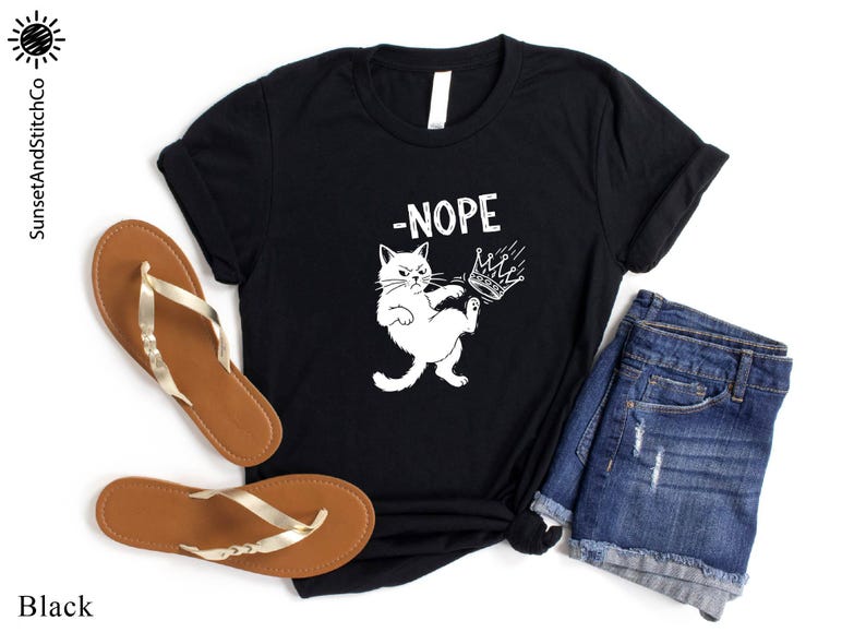May include: Black t-shirt featuring a white cat graphic kicking a crown, with the word "-NOPE" above. The shirt is styled with denim shorts and brown sandals with gold straps. The brand name "SunsetAndStitchCo" is visible.