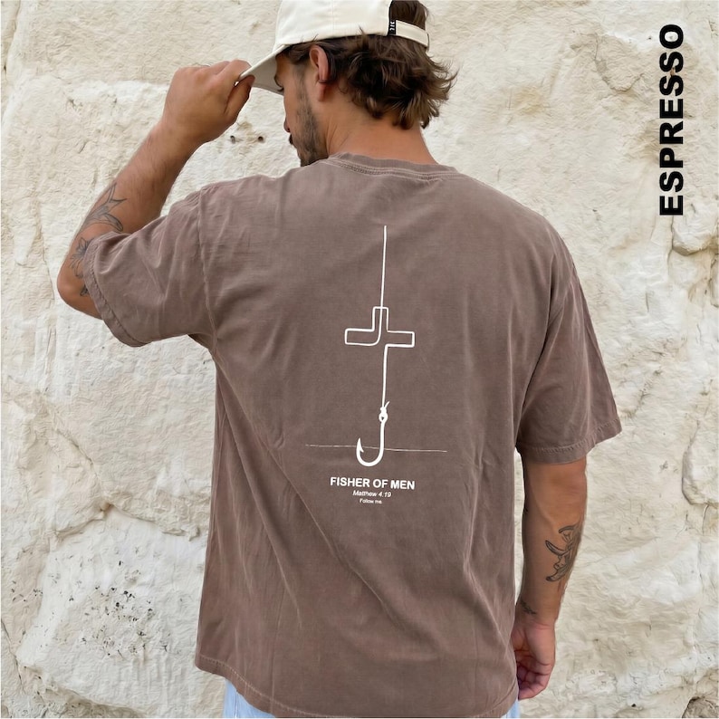May include: A brown short-sleeve t-shirt featuring a white line art graphic on the back. The design depicts a fishing hook shaped like a cross with the text "FISHER OF MEN" and "Matthew 4:19" printed below it. The word "ESPRESSO" is printed vertically.