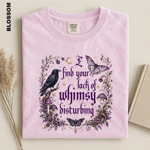 Puede incluir: Una camiseta rosa claro con un estampado gr&aacute;fico que muestra un cuervo negro, setas moradas, mariposas y el texto I find your lack of whimsy disturbing. La camiseta tiene cuello redondo y una etiqueta de la marca Comfort Colors en el interior.