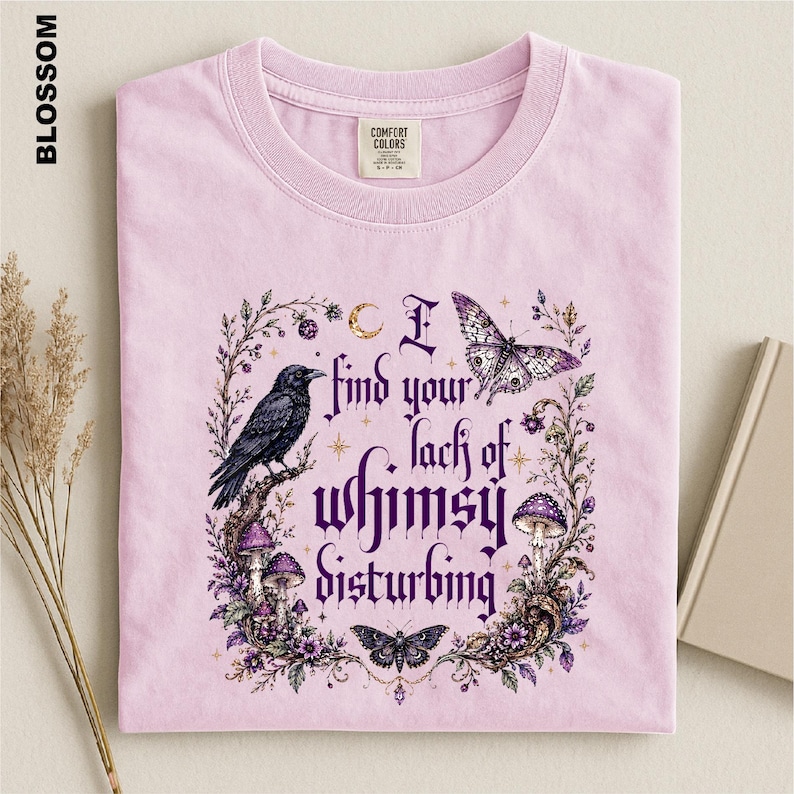Puede incluir: Una camiseta rosa claro con un estampado gr&aacute;fico que muestra un cuervo negro, setas moradas, mariposas y el texto I find your lack of whimsy disturbing. La camiseta tiene cuello redondo y una etiqueta de la marca Comfort Colors en el interior.