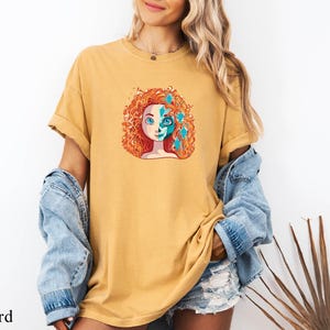 A woman wearing a yellow t-shirt with a colorful graphic of a woman's face and a denim jacket stands in front of a white wall.