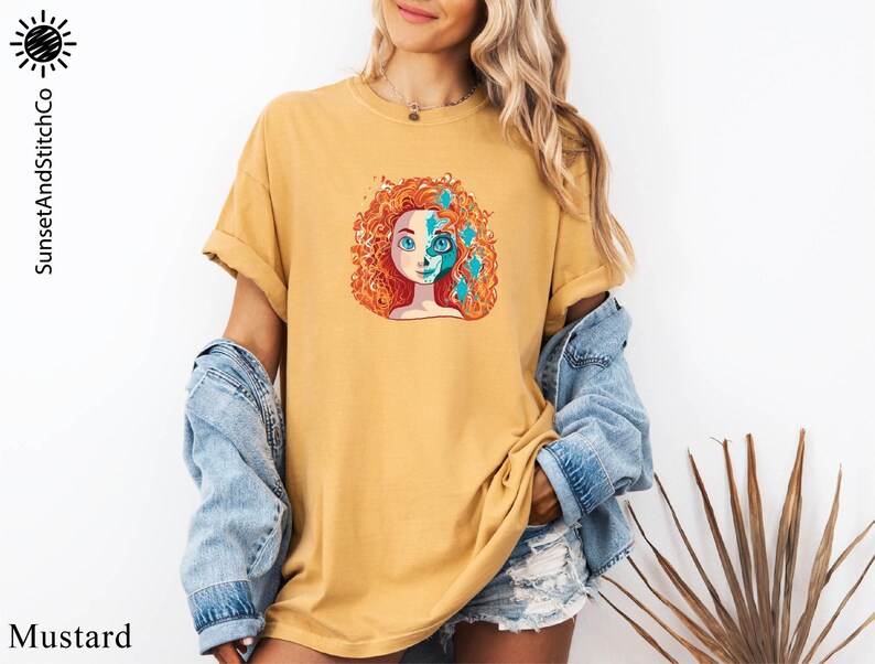A woman wearing a yellow t-shirt with a colorful graphic of a woman's face and a denim jacket stands in front of a white wall.