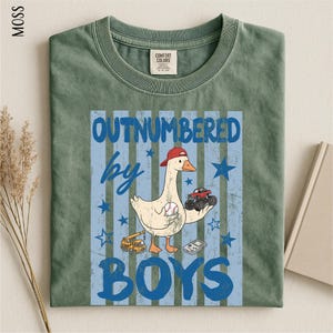 A green t-shirt with a graphic design featuring a cartoon duck holding a baseball and a truck, along with the text "OUTNUMBERED BY BOYS".