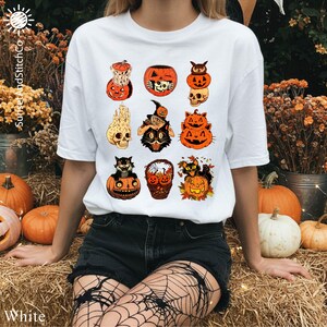 A woman wearing a white t-shirt with a graphic of Halloween-themed cats and pumpkins, surrounded by pumpkins, flowers, and a spooky atmosphere.