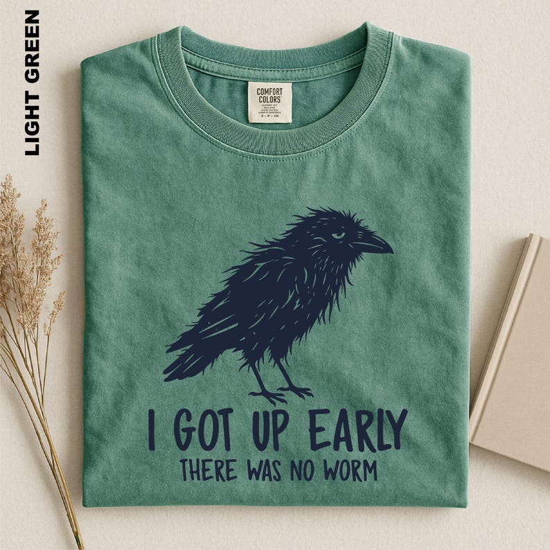 May include: A light green crew neck t-shirt featuring a dark navy blue graphic of a ruffled crow. Below the bird, the text reads: I GOT UP EARLY THERE WAS NO WORM. The shirt has a Comfort Colors brand label on the inside of the collar.