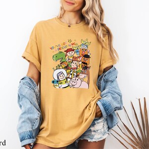 A woman wearing a yellow t-shirt with a colorful graphic of various cartoon characters, including a pig, a dinosaur, and a robot. She is also wearing a denim jacket and denim shorts.