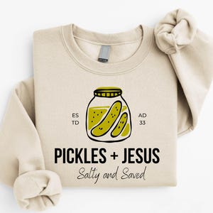 A beige sweatshirt with a graphic design featuring a jar of pickles and the text "Pickles + Jesus Salty and Saved" printed on it.
