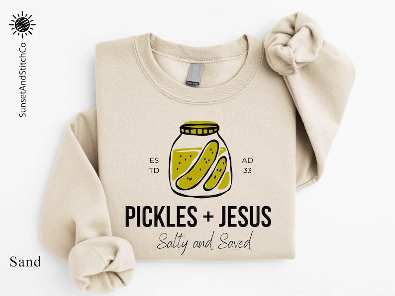 A beige sweatshirt with a graphic design featuring a jar of pickles and the text "Pickles + Jesus Salty and Saved" printed on it.