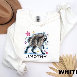 May include: A white long-sleeve sweatshirt featuring a graphic illustration of a raccoon with shaggy fur. The design includes colorful hand-drawn style stars and the name JIMOTHY printed in black capital letters below the raccoon character.