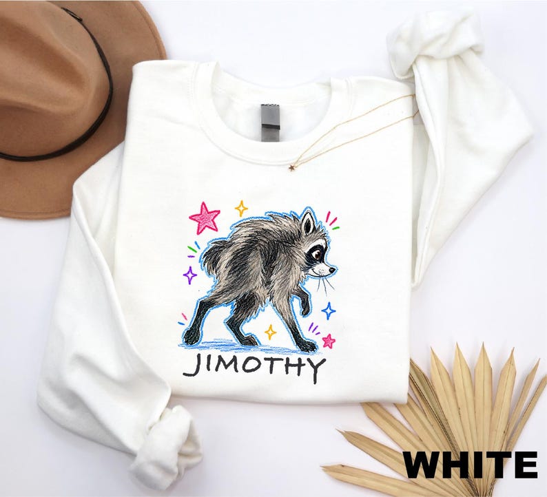 May include: A white long-sleeve sweatshirt featuring a graphic illustration of a raccoon with shaggy fur. The design includes colorful hand-drawn style stars and the name JIMOTHY printed in black capital letters below the raccoon character.