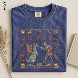 May include: A navy blue crew neck t-shirt featuring a graphic print of a medieval knight touching the nose of a green dragon. The design includes the text LE BOOP, surrounded by a decorative rectangular border with floral and mythical motifs.