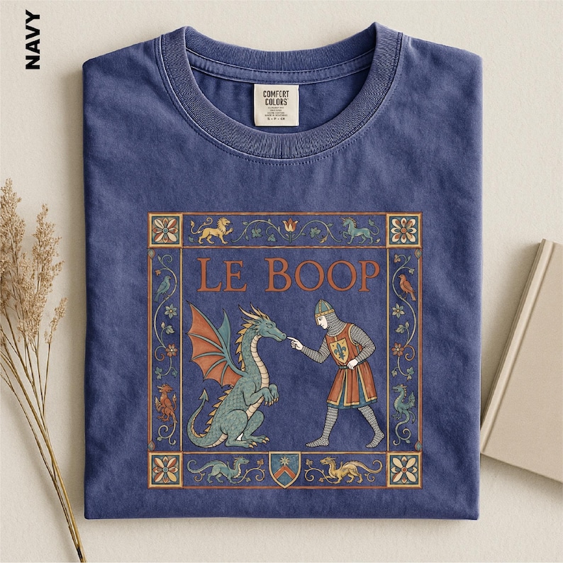 May include: A navy blue crew neck t-shirt featuring a graphic print of a medieval knight touching the nose of a green dragon. The design includes the text LE BOOP, surrounded by a decorative rectangular border with floral and mythical motifs.