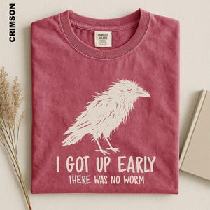 May include: A crimson short-sleeved t-shirt featuring a white graphic of a bird and the text I GOT UP EARLY THERE WAS NO WORM. The shirt has a crew neckline and a Comfort Colors brand label on the inside collar. The fabric has a washed appearance.