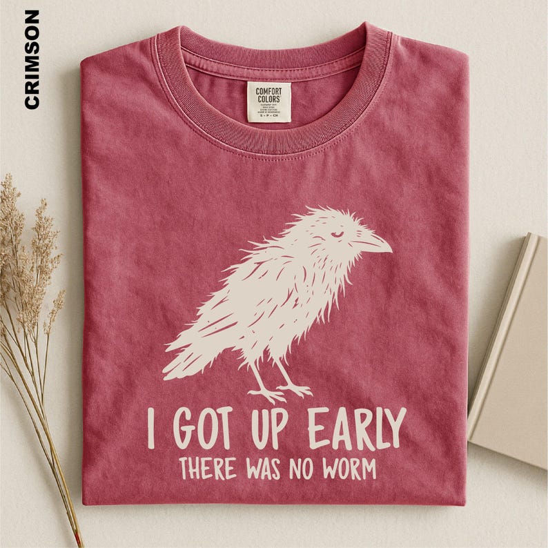 May include: A crimson short-sleeved t-shirt featuring a white graphic of a bird and the text I GOT UP EARLY THERE WAS NO WORM. The shirt has a crew neckline and a Comfort Colors brand label on the inside collar. The fabric has a washed appearance.